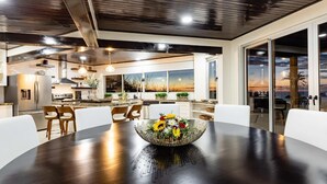 A modern kitchen with stainless steel appliances, a dining area with a dark wooden table and white chairs, and large windows offering a view of the sunset. - Playa Hermosa Villa • 4BR • Infinity Pool • Ocean & Bay View • Recently Renovated • Sunset Ridge (Playa Hermosa)