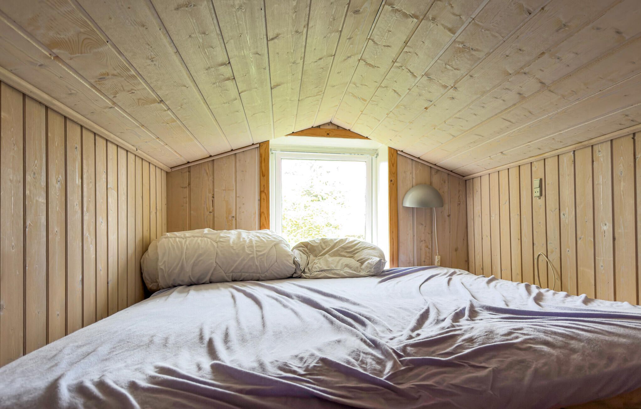 Ein Schlafzimmer mit schrägverdeckter Decke, Holzwänden, einem Bett mit Kissen, einer Lampe und einem Fenster.