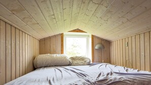 A bedroom with a sloped ceiling, wooden walls, a bed with pillows, a lamp, and a window.