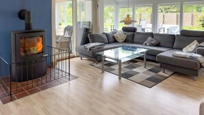 A modern living room with a wood-burning stove, grey sofa, glass coffee table, and a view of the garden through large windows.