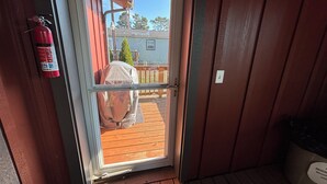 Une porte-fenêtre coulissante donne sur une terrasse en bois équipée d’un barbecue couvert, d’un extincteur fixé au mur et d’une poubelle de recyclage.