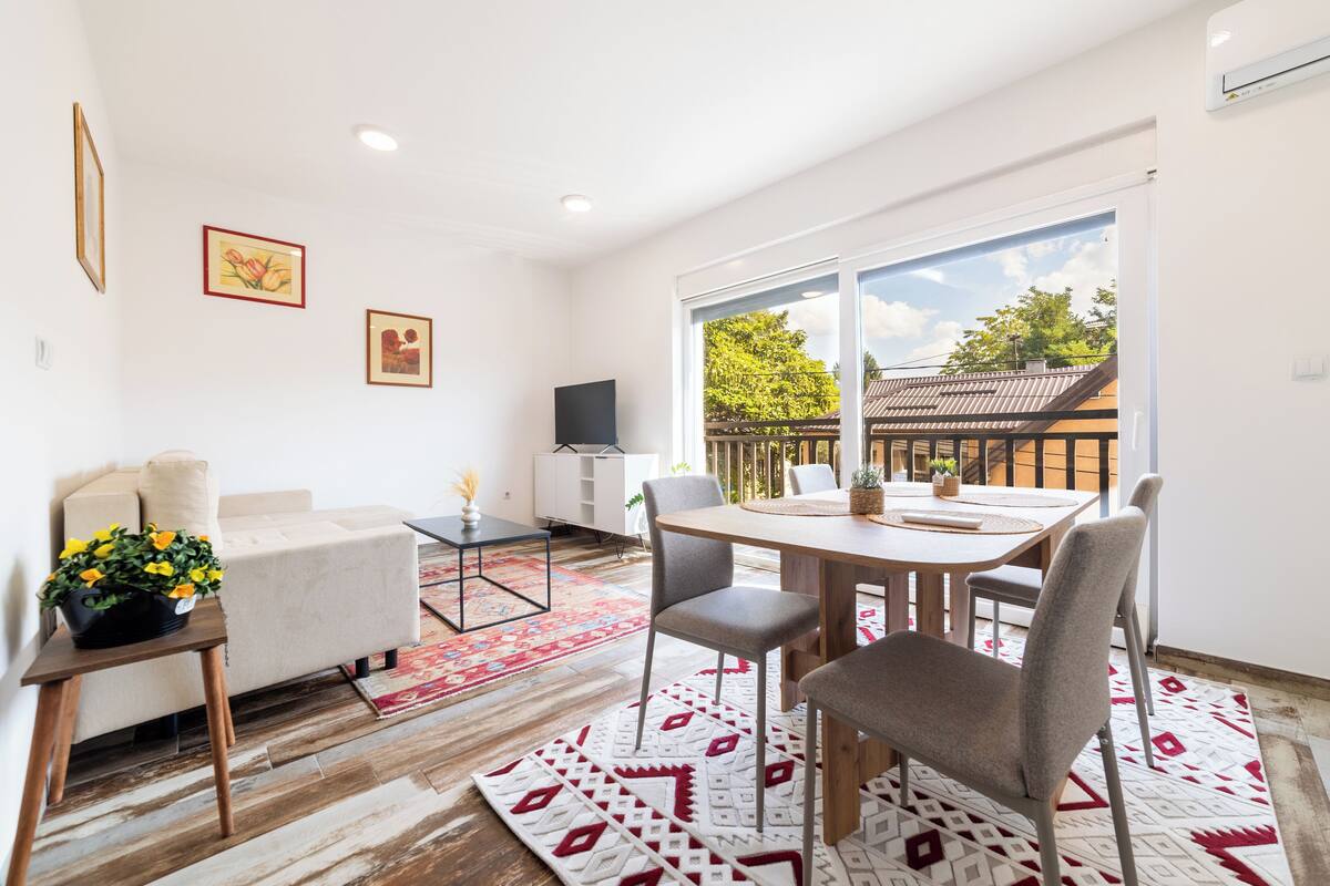 Una sala de estar moderna con sofá, mesa de comedor y sillas, televisor y ventana grande con vistas a un edificio y árboles.