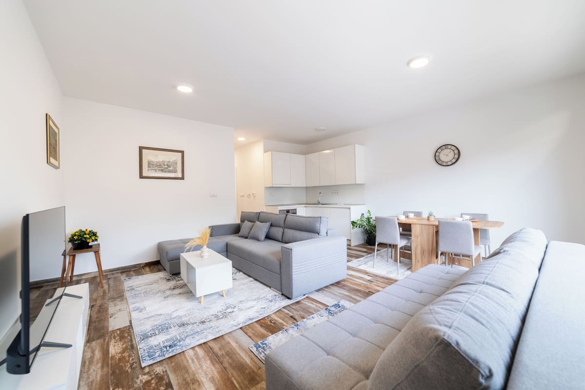 Una sala de estar moderna con un sofá gris, una mesa de centro blanca y un televisor. Hay una zona de comedor con una mesa de madera y sillas.
