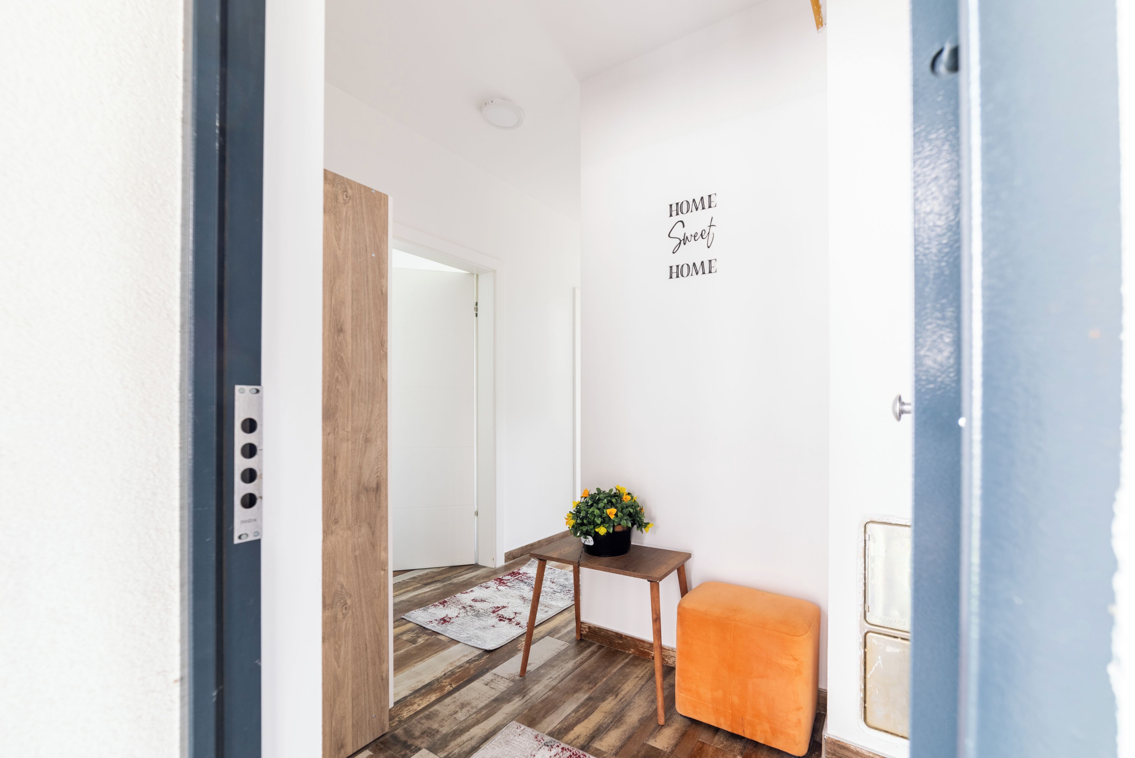 A hallway with a wooden floor, a small table with a potted plant, a bench, and a wall sign that says "HOME Sweet HOME".