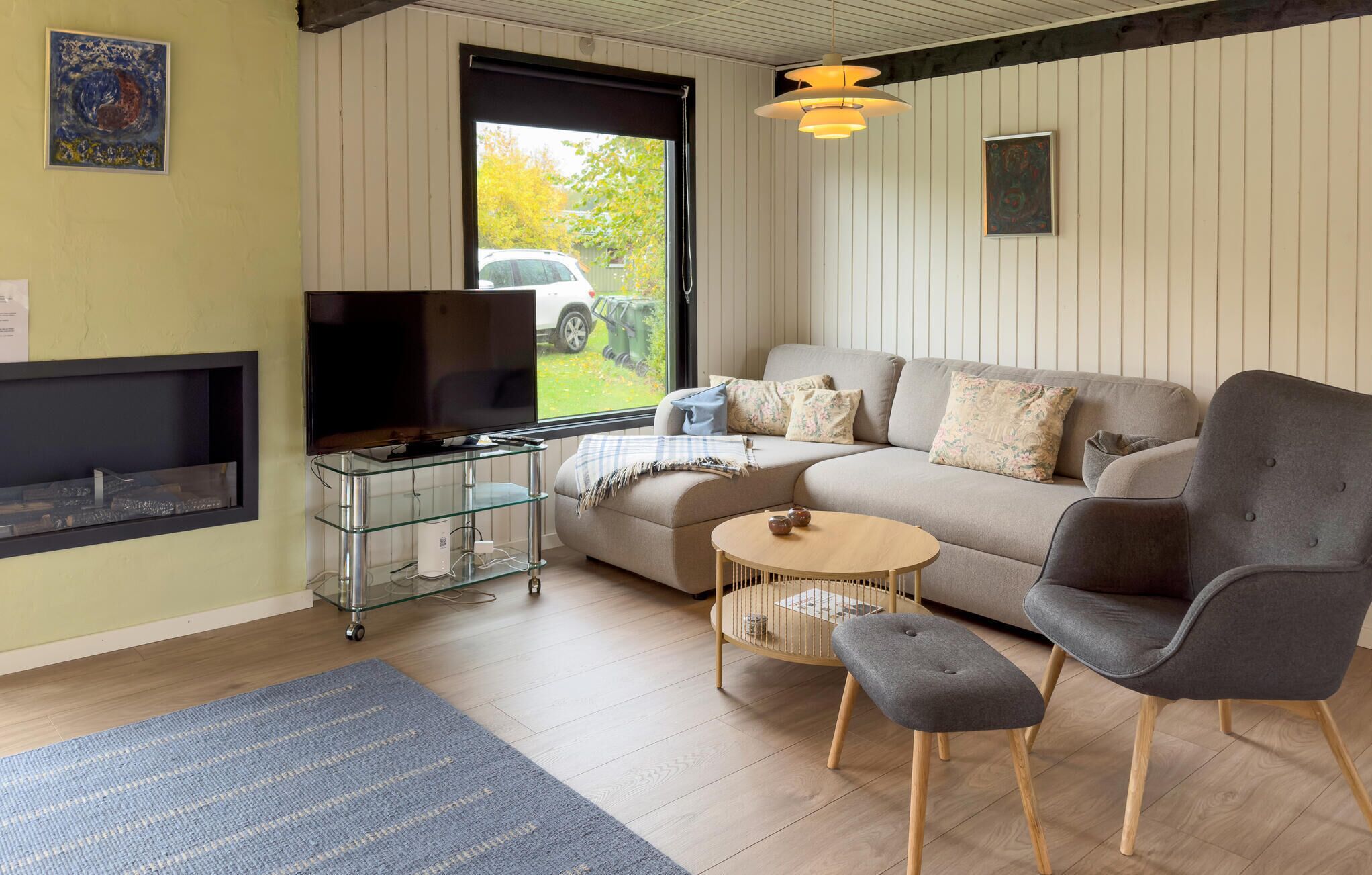 Een woonkamer met een bank, twee stoelen, een tv op een glazen standaard, een kleine tafel en een raam met uitzicht op een auto en groen.