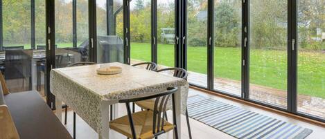 A conservatory with a table and chairs, large glass doors, and a view of a green garden.