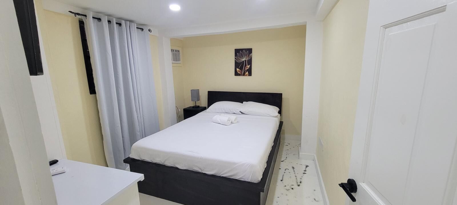 A small, neatly arranged bedroom with a single bed, a bedside table, a lamp, and a wall-mounted air conditioning unit.
