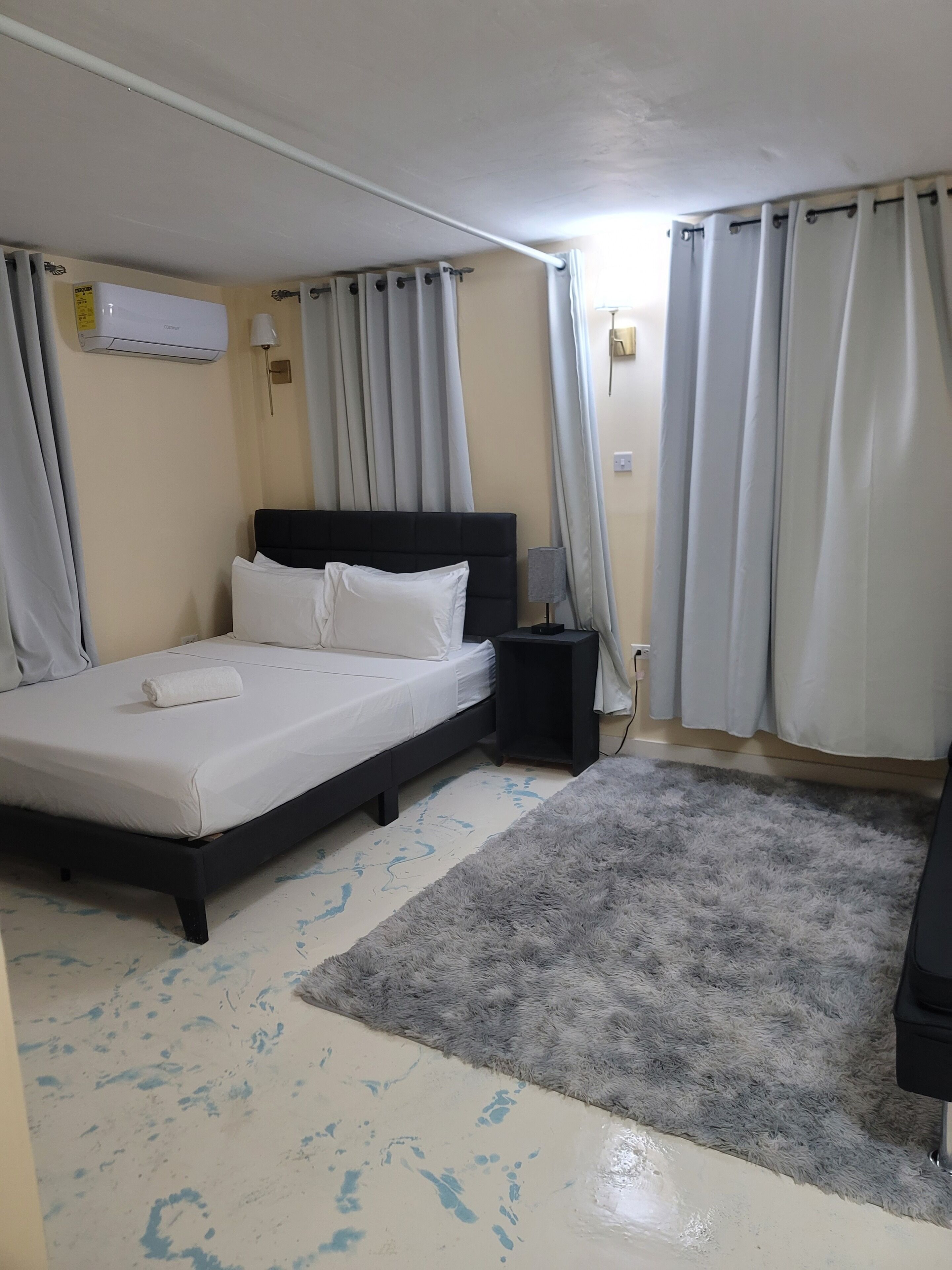 Una camera d'albergo con un letto, un comodino, un condizionatore d'aria, una lampada e un tappeto.