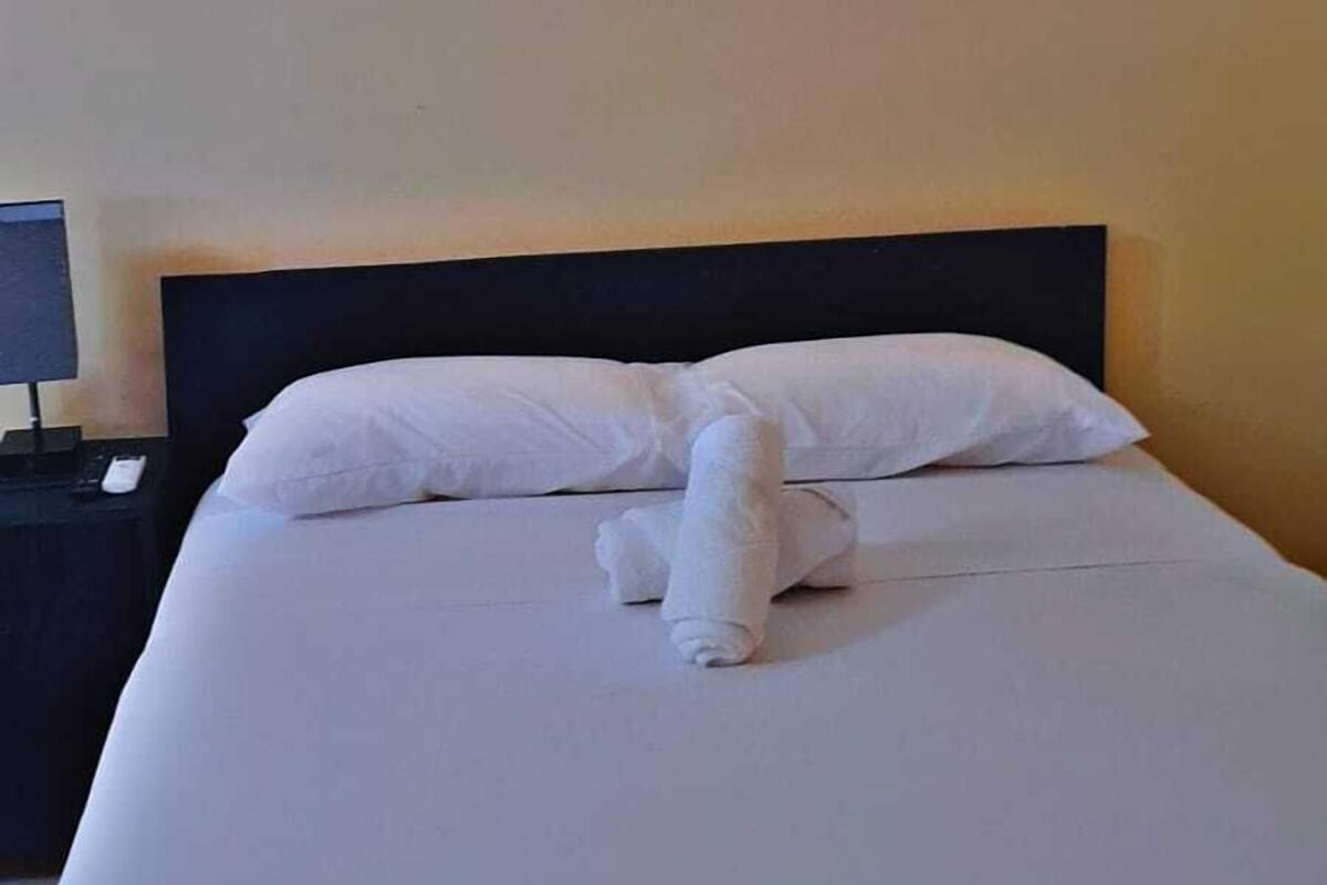A neatly made bed with white linens and pillows, a bedside table with a lamp, and a wall-mounted light fixture.
