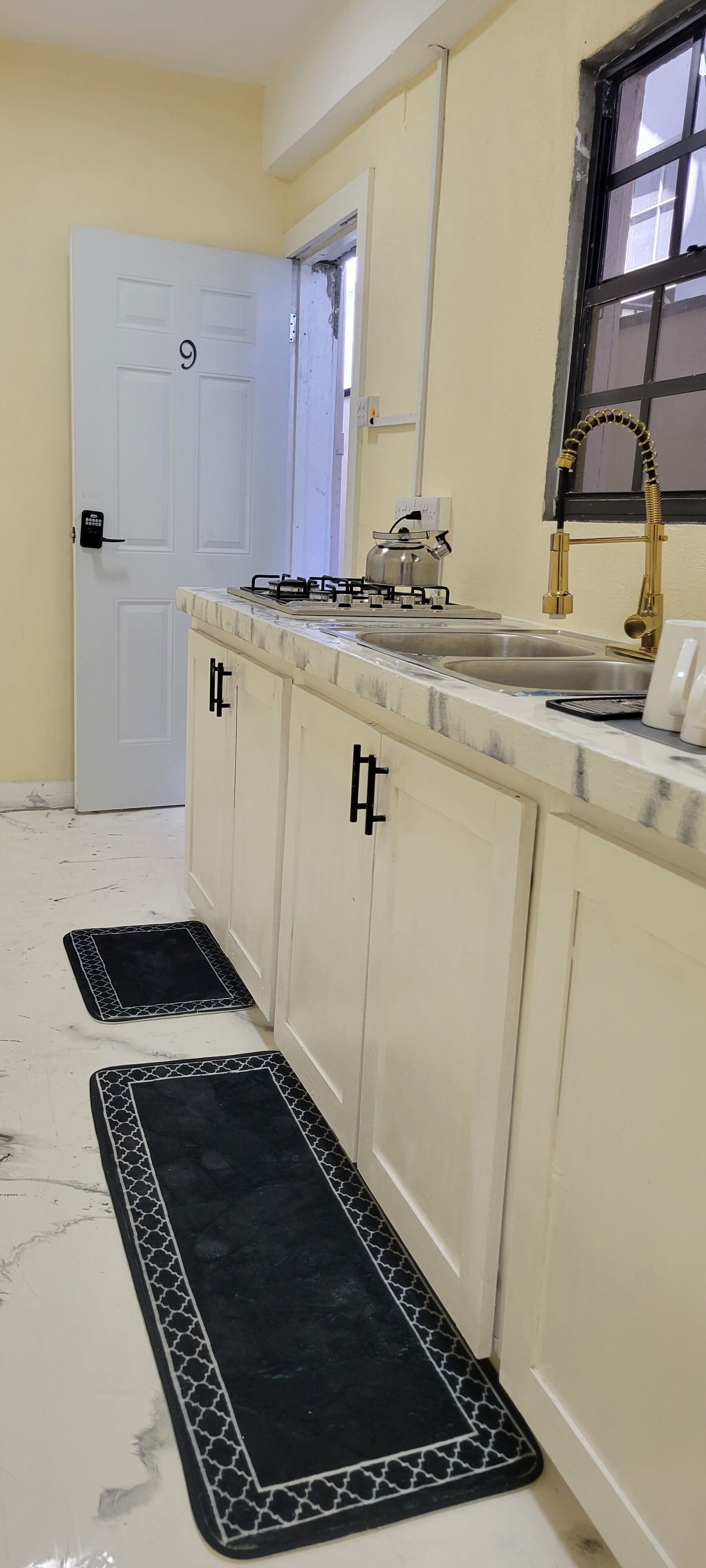 A kitchen with a white door marked with the number 9, a gas stove, a sink with a gold faucet, and black and white patterned mats on the floor.