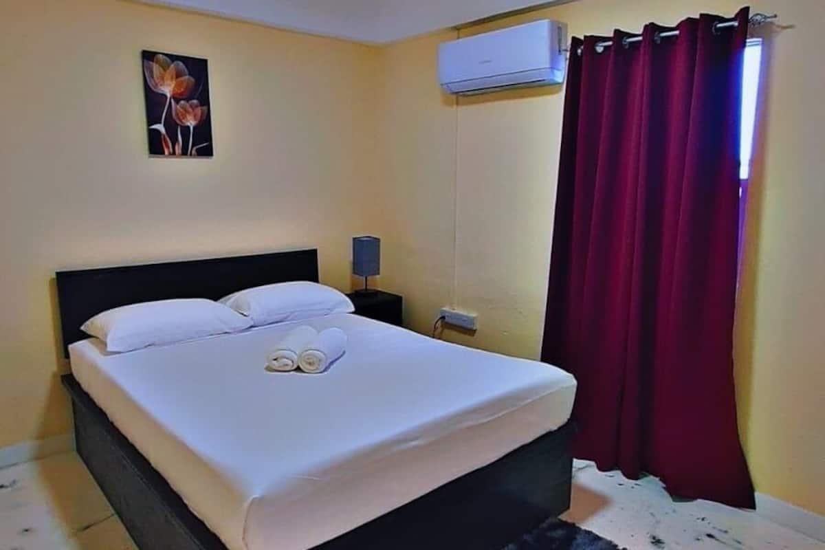 A bedroom with a bed, a wall-mounted air conditioner, a dark rug, and a painting.