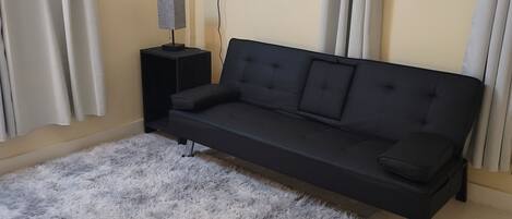 Un soggiorno moderno con un divano nero, un tappeto grigio soffice, un piccolo tavolino nero con una lampada e un televisore montato a parete.