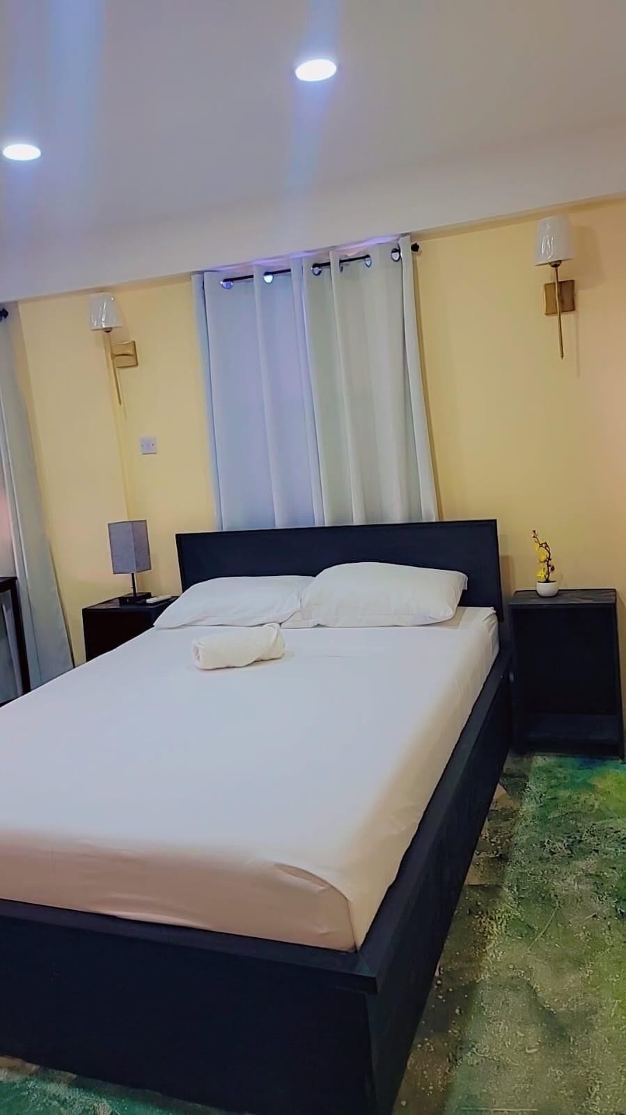 A hotel room with a bed, a nightstand, a lamp, and a wall-mounted light fixture.