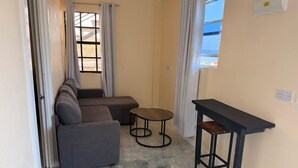 A living room with a sofa, a small round table, a black bench, and a window with curtains. - Oceanview Suites (St. George's)
