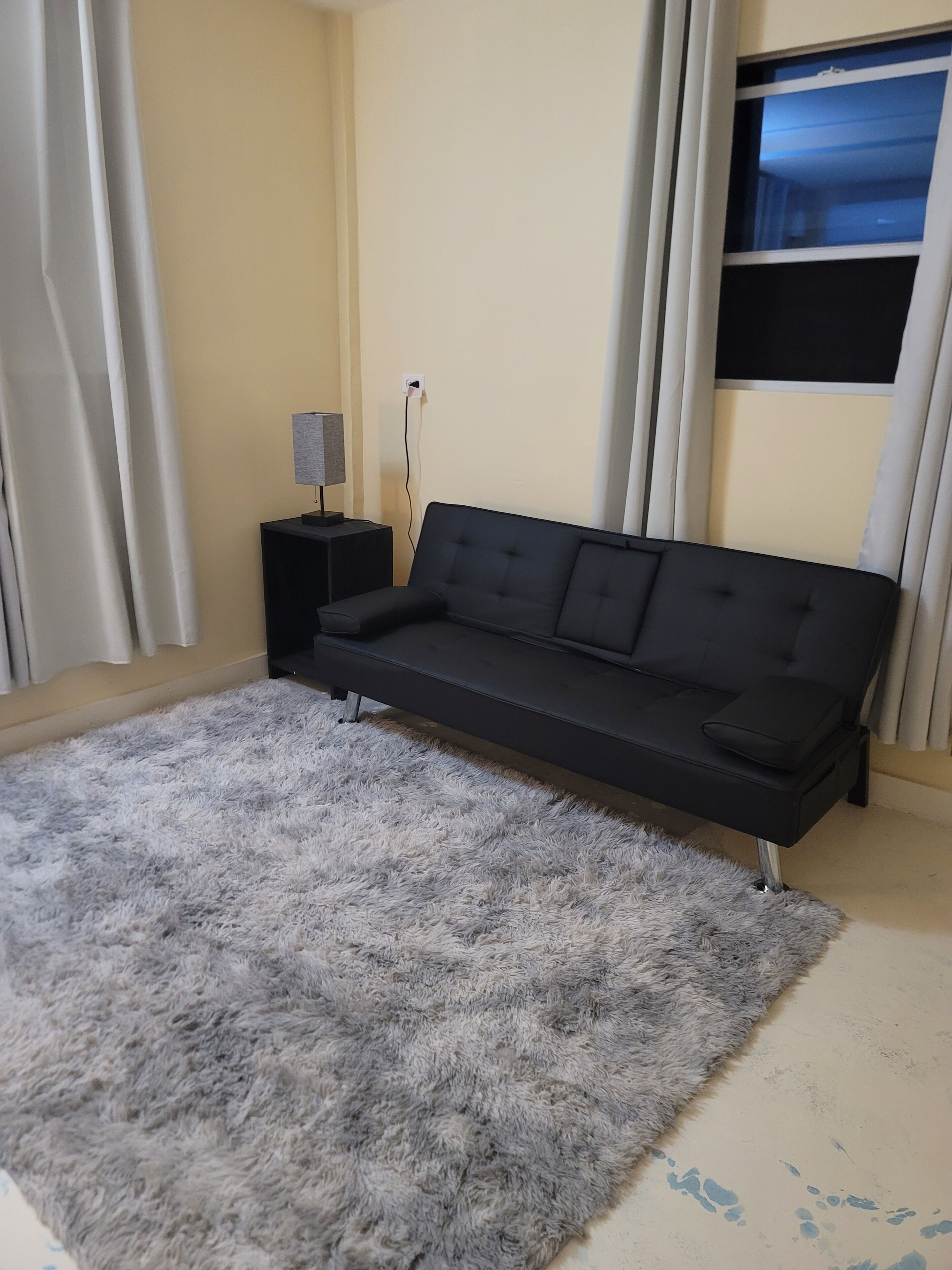 A modern living room with a black sofa, a fluffy grey rug, a small black side table with a lamp, and a TV mounted on the wall.