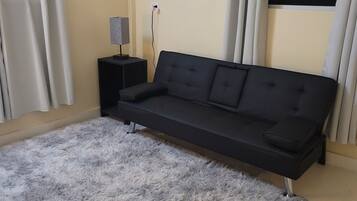 A modern living room with a black sofa, a fluffy grey rug, a small black side table with a lamp, and a TV mounted on the wall.