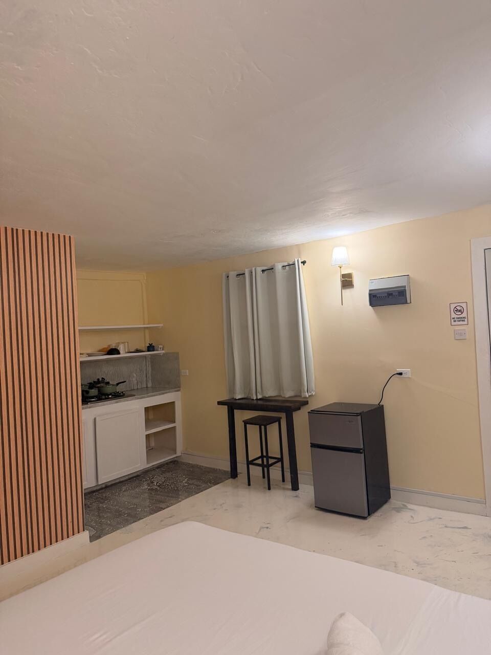 A compact room with a bed, a kitchenette, a small table, a refrigerator, and a wall-mounted light fixture.
