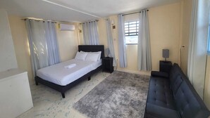 A bedroom with a bed, a sofa, a nightstand, an air conditioning unit, and a window with curtains. - Oceanview Suites (St. George's)