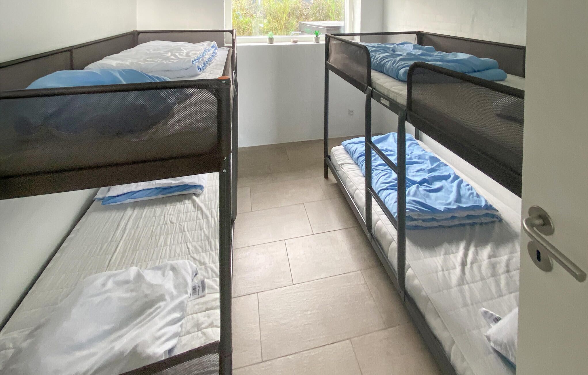 A dormitory room with bunk beds, a window with a view of greenery, and a door with a handle.