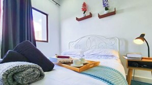 Un lit bien fait, avec une tablette sur laquelle sont posés des livres, une tasse et un mug, une lampe de chevet et une fenêtre avec des rideaux.