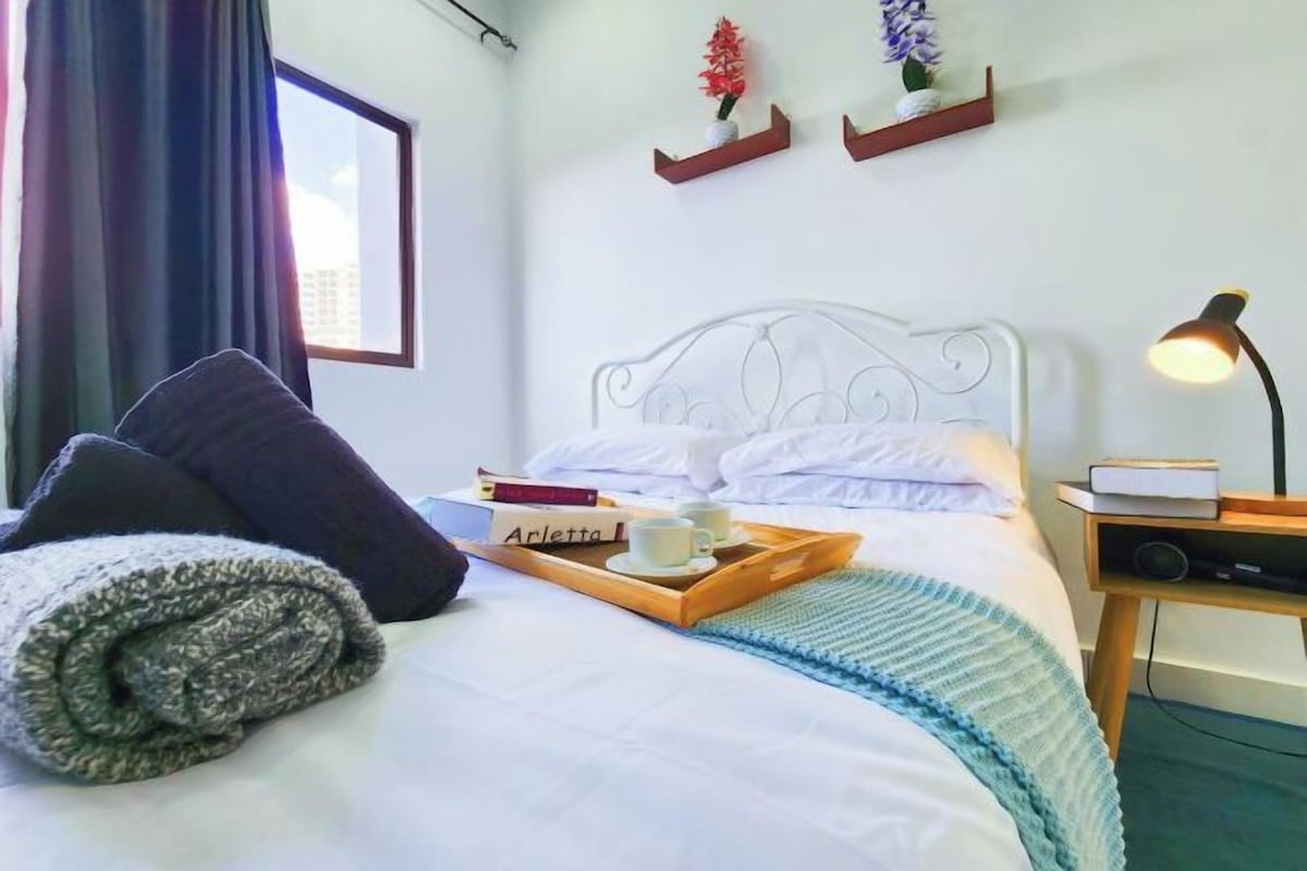 A neatly made bed with pillows, a bedside table with a lamp, a tray with a book and a cup, and a window with curtains.