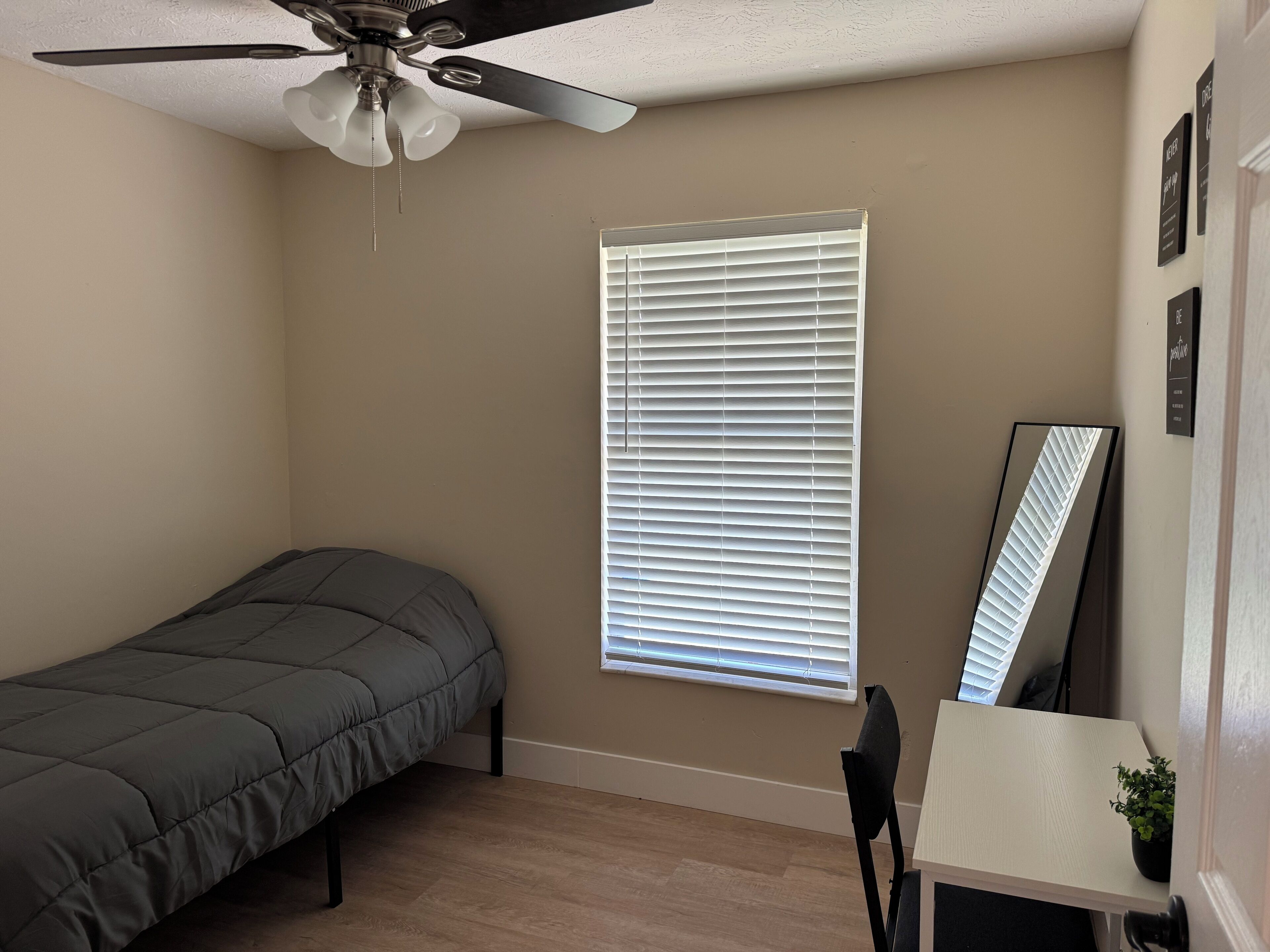 A small, neatly arranged bedroom with a single bed, a ceiling fan, a window with blinds, a small table with a plant, and a mirror.