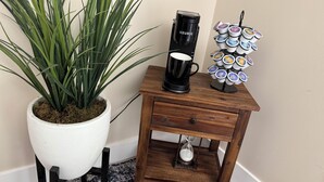Uma pequena mesa de apoio de madeira com uma cafeteira preta, uma caneca e uma seleção de cápsulas de café.