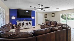 Uma sala de estar com sofá seccional, TV de tela plana, ventilador de teto e um armário com objetos decorativos.