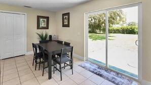 Uma área de refeições com mesa, cadeiras, um vaso com planta e uma porta de vidro deslizante para um quintal.