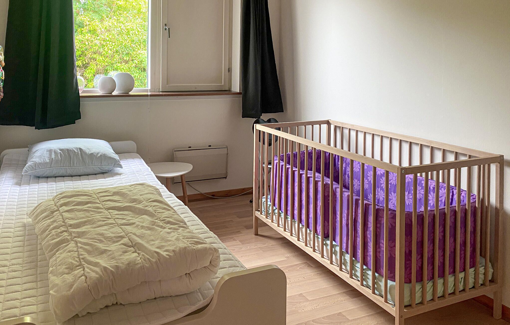 A bedroom with a bed, a crib, a radiator, and a window with curtains.