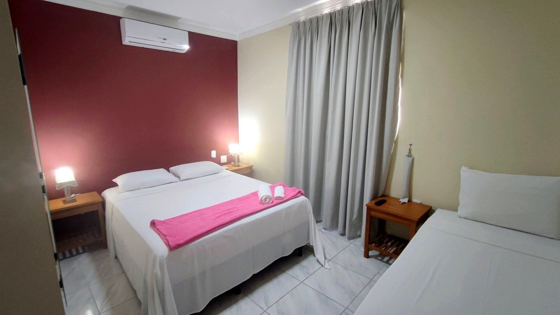A hotel room with a bed, two bedside tables, a nightstand, a lamp, a window with curtains, and an air conditioning unit.