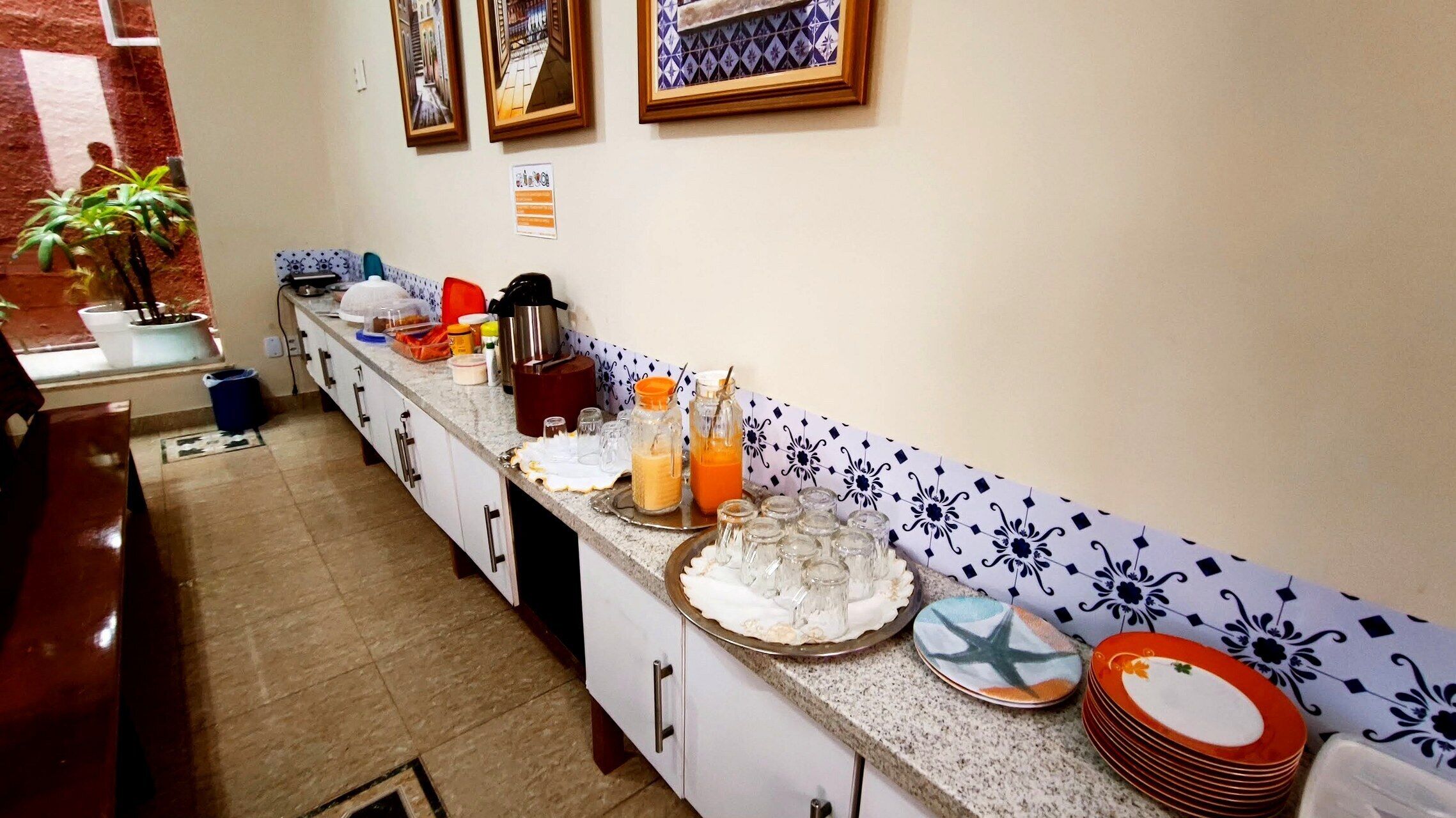 Un área de cocina con encimera de mármol, cafetera y diversos alimentos. Hay plantas en macetas y un salpicadero de azulejos.