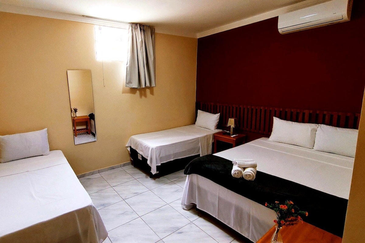 A hotel room with two single beds, a small table with a lamp, a mirror, and a radiator.