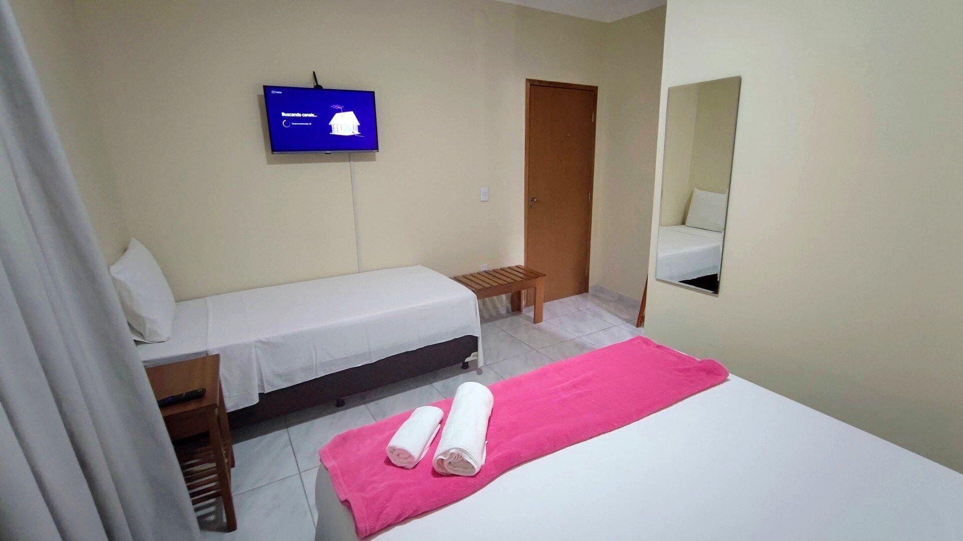 A small, clean room with a bed, a bench, a TV, and a mirror.