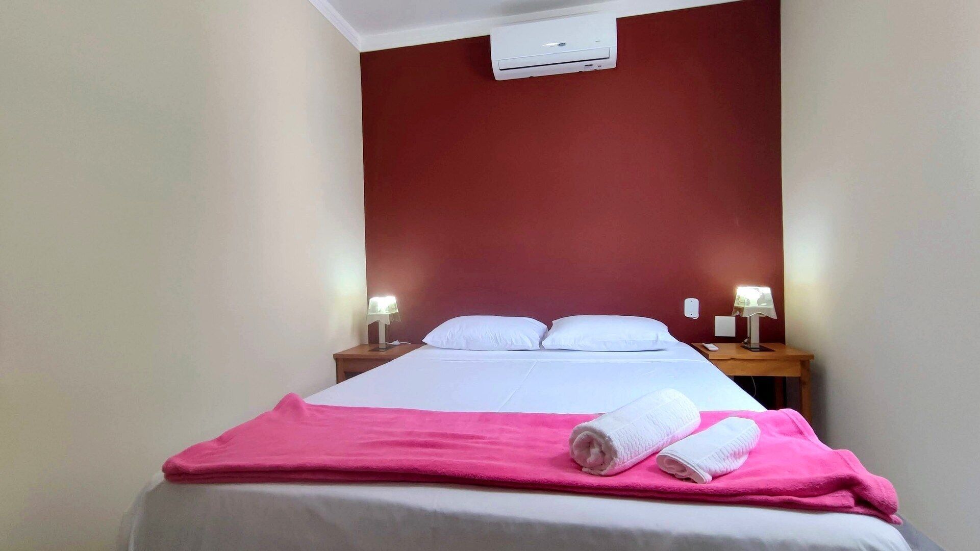 Habitación de hotel con cama, dos mesitas de noche con lámparas, una pared de acento roja y un equipo de aire acondicionado.