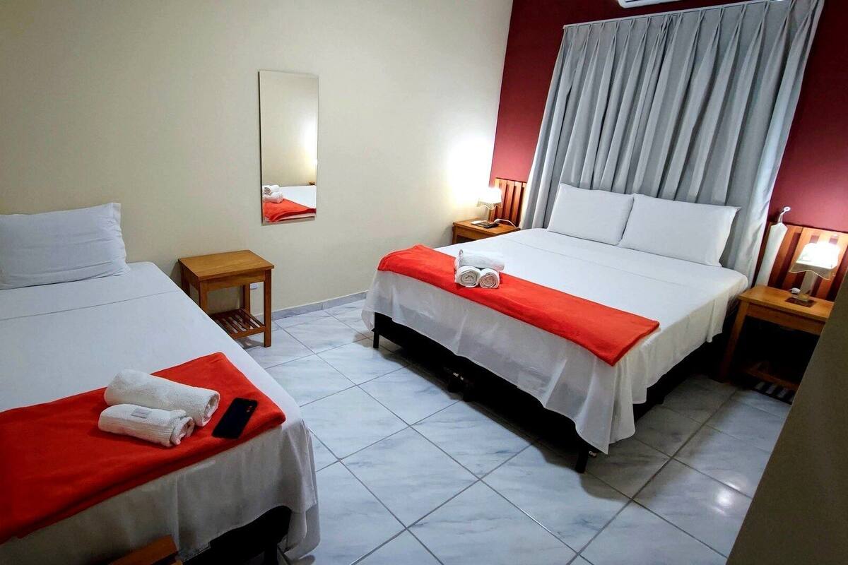 A hotel room with two beds, a small table, a mirror, and a window with curtains.