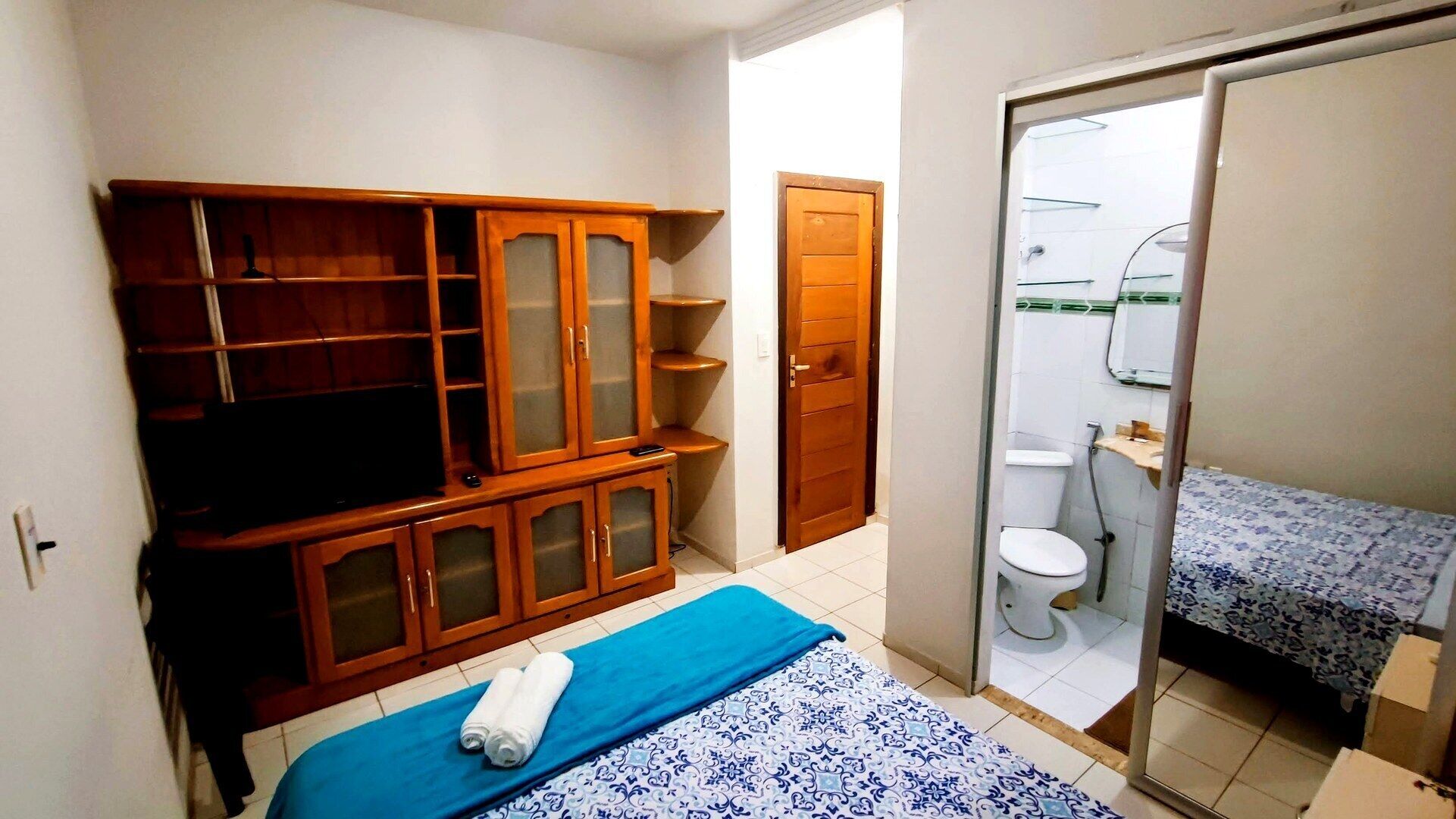 A compact room with a wooden cabinet, a bed with a blue and white patterned bedspread, a white toilet, and a small bathroom with a mirror and towel rack.
