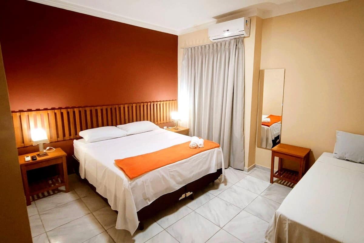 A hotel room with a bed, a bedside table, a nightstand, a mirror, and an air conditioning unit.