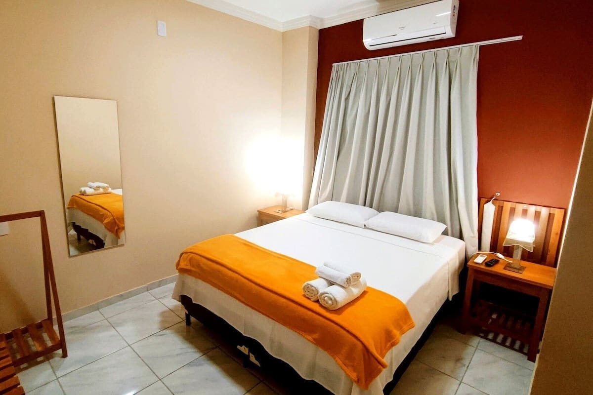 A hotel room with a bed, a bedside table with a lamp, a mirror, and an air conditioning unit.