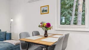 Ein modernes Zimmer mit einem Holztisch, grau gepolsterten Stühlen, einer Blumenvase, einem gerahmten Bild an der Wand und einem Fenster mit Blick auf Bäume.