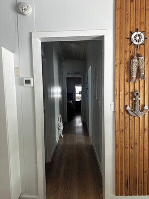 Een smalle gang met houten vloer, witte muren en een houten paneel aan de rechterkant. Een witte deur bevindt zich aan de linkerzijde, en er is een wandlamp te zien.