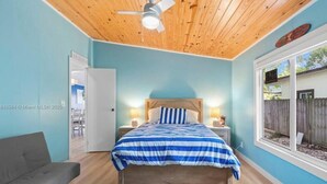 A bedroom with a wooden ceiling, a bed with a blue and white striped bedspread, a gray sofa, and a window with a view of the outside.