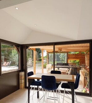 Un espace repas moderne avec une table en bois et des chaises bleues, de grandes fenêtres et une cuisine extérieure visible à travers la vitre.