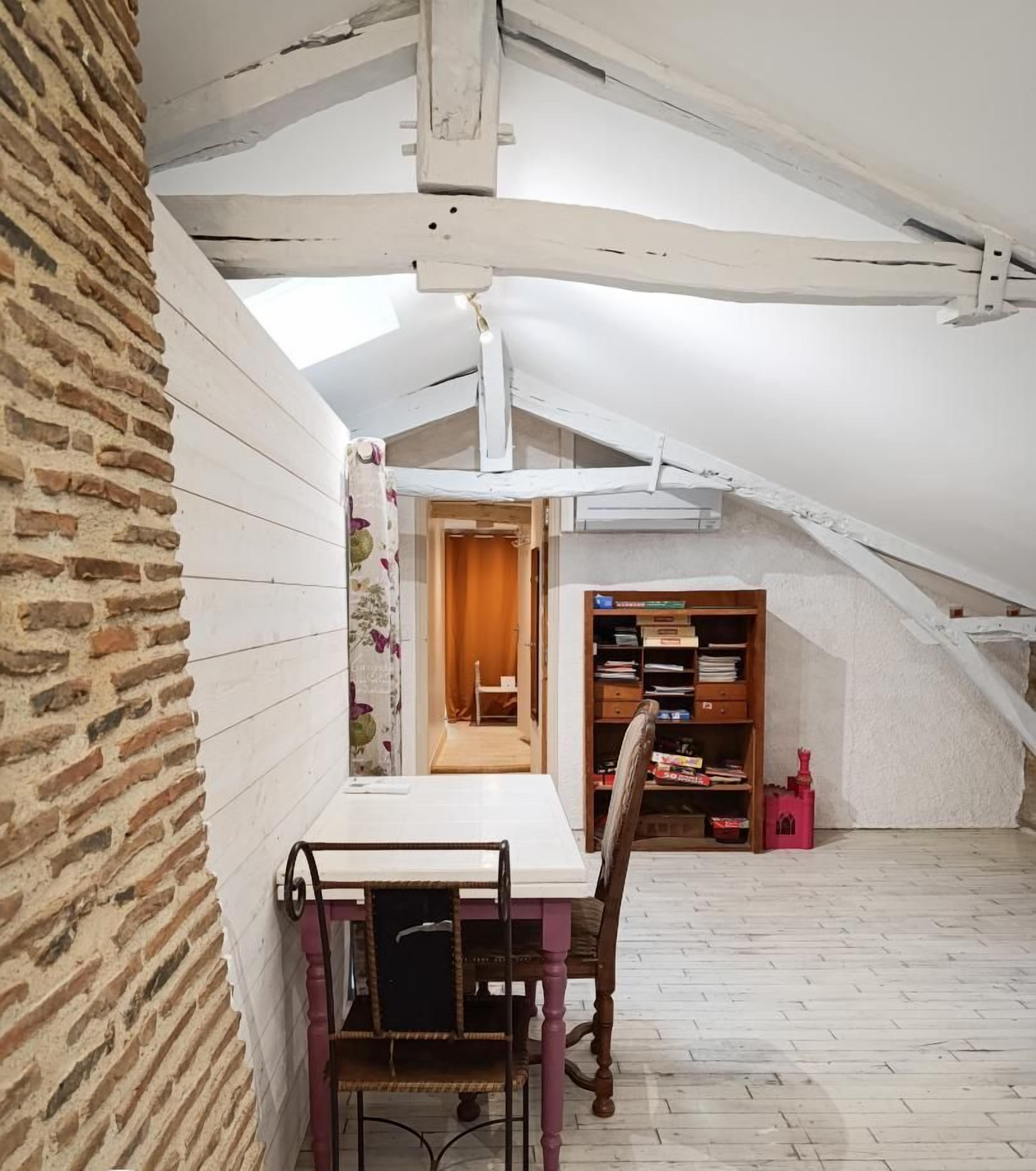 A room with a sloped ceiling, a brick wall, a table with chairs, and a bookshelf.