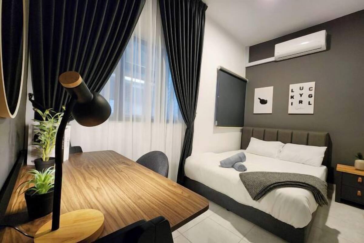 Una habitació d'hotel moderna amb escriptori de fusta, llit amb roba blanca, làmpada negra i aire condicionat fixat a la paret.