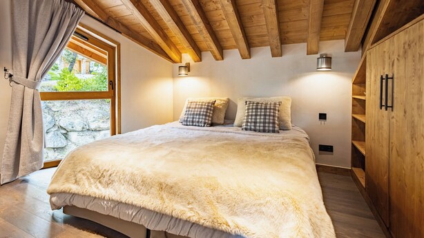 A bedroom with a large bed, wooden ceiling beams, a window with curtains, and a stone wall visible through the window. - Chalets Killington #4 - 4 guests (Les Allues)