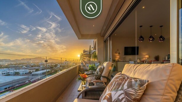 A balcony with a sofa and chairs, a TV, and a view of the cityscape at sunset. - New 2 BR Penthouse Overlooking the Sea of Cortez (Cabo San Lucas)