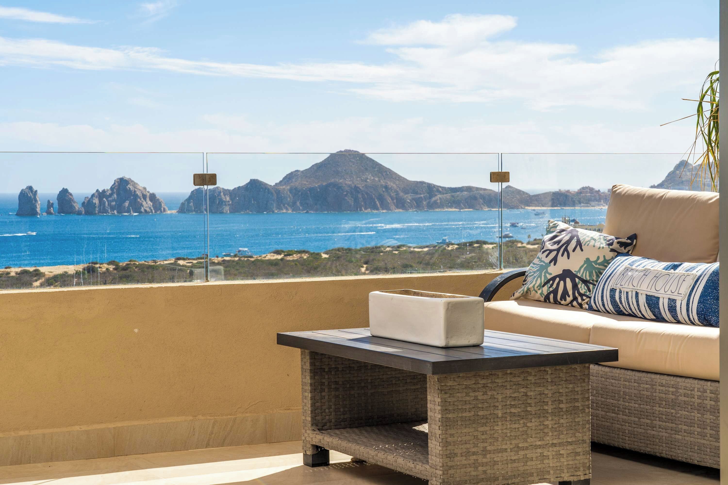 A balcony with a view of the sea and mountains, a sofa with a cushion, and a small table with a box on it.