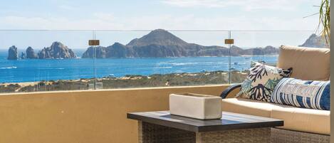 A balcony with a view of the sea and mountains, a sofa with a cushion, and a small table with a box on it.