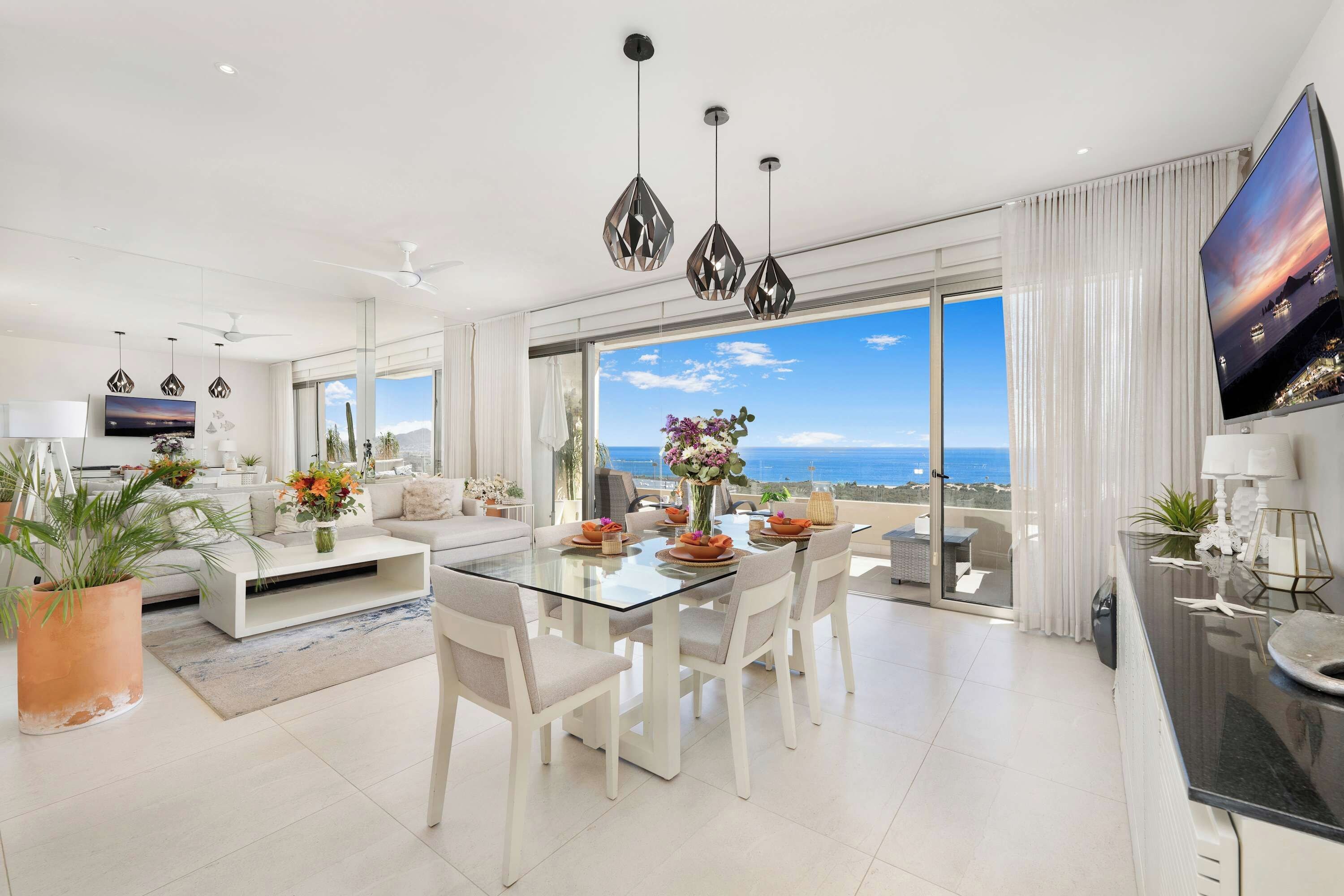 A modern living room with a glass dining table, white sofas, and a large TV screen.